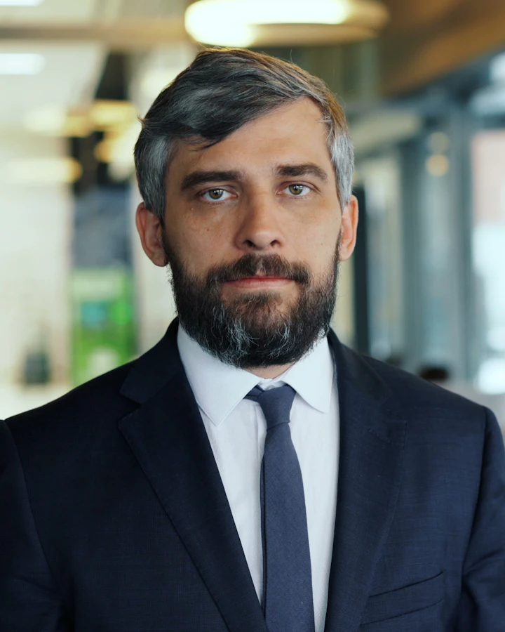 Homem de barba grisalha, terno e gravata, olhando direto para a câmera